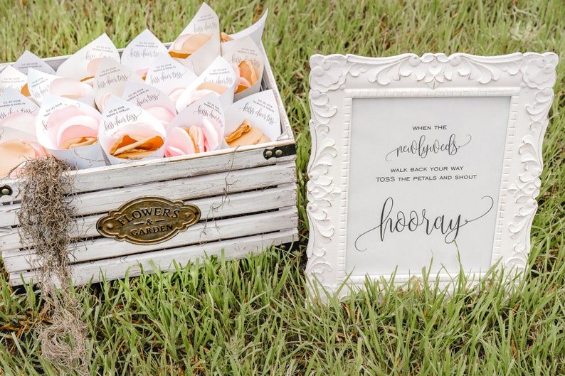May include: A white wooden box filled with small white cones containing pink flower petals. A white sign with black calligraphy reads "When the newlyweds walk back your way toss the petals and shout hooray".
