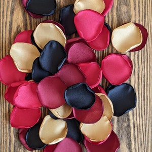 Może przedstawiać: Stosik czarnych, bordowych i złotych satynowych płatków róż. Płatki są ułożone w przypadkowym wzorze i rozrzucone na drewnianej powierzchni.