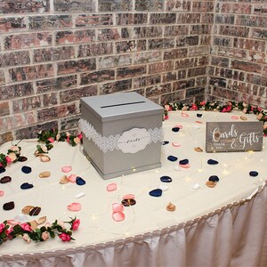 May include: A silver wedding card box with a white lace trim and a sign that says "Cards & Gifts" with a thank you note. The box is on a table with a white tablecloth and is surrounded by pink and blue flower petals.