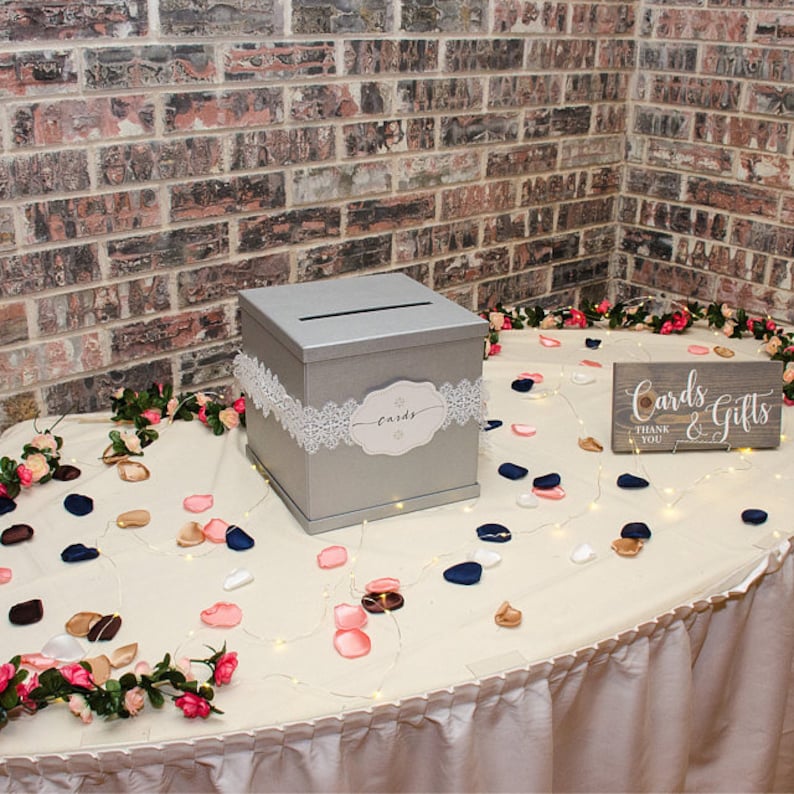 May include: A silver wedding card box with a white lace trim and a sign that says "Cards & Gifts" with a thank you note. The box is on a table with a white tablecloth and is surrounded by pink and blue flower petals.