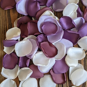 Könnte beinhalten: Ein Haufen weißer und lila Satin-Blütenblätter. Die Blütenblätter sind in einem zufälligen Muster angeordnet und auf einer Holzoberfläche verstreut.