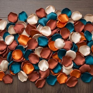 May include: A collection of artificial flower petals in a mix of satin-finish colors including deep teal, burnt orange, rust brown, and cream. The petals are scattered in a dense, overlapping arrangement, showcasing their smooth, rounded shapes.