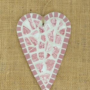 May include: A pink and white heart-shaped mosaic made from broken pieces of china. The mosaic is hanging on a burlap background.