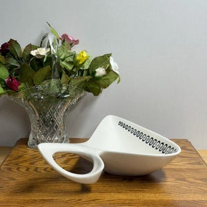 May include: A white ceramic serving dish with a handle and a black zigzag pattern along the rim. The dish is on a wooden surface, with a glass vase of flowers in the background.