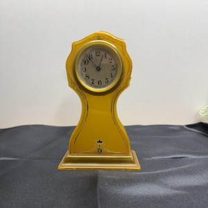May include: A vintage yellow mantel clock with a round clock face and black numerals. The clock has a decorative, ornate design with gold accents and a rectangular base. The clock is a decorative item.