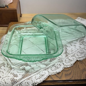 Könnte beinhalten: Eine quadratische, hellgrüne Butterdose aus Glas mit Deckel. Die Dose hat einen dekorativen, verzierten Rand und einen Griff am Deckel. Die Dose steht auf einer weißen Spitzendecke.