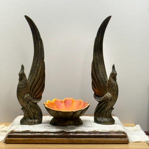 May include: Two bronze-colored phoenix sculptures flank a ceramic bowl with a flower-like design. The bowl features a vibrant orange and red interior. The sculptures and bowl are displayed on a wooden base.