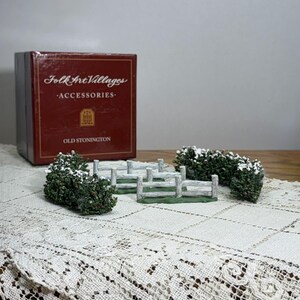 May include: A Folk Art Villages accessory set featuring miniature fences and bushes with snow. The set includes a red box with gold lettering that reads "Folk Art Villages Accessories Old Stonington."