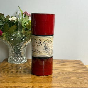 May include: A three-sectioned, cylindrical container with a red lacquer finish. The middle section features a cream-colored band with a black and white illustration of a bird and foliage. The container is displayed on a wooden surface.