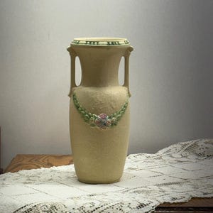 May include: A beige ceramic vase with two handles and a decorative floral garland. The vase has a textured surface and a green and cream-colored rim. The floral garland features pink, blue, and green accents.