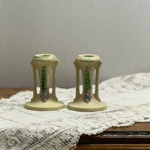 May include: Two antique candle holders. The candle holders are a pale yellow with green and blue floral accents. The design includes a floral motif and vertical supports. The candle holders are displayed on a white lace tablecloth.