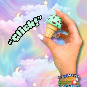 May include: A small ice cream cone charm with mint-chocolate chip ice cream and a waffle cone is held up. The charm is on a light blue beaded chain. The background is a pastel sky with clouds, stars, and moons. The text "Click!" is in the upper left.