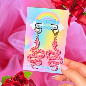 May include: Pink snake earrings with red heart details, hanging from gold hoops. The earrings are displayed on a card with the text "cosmic love" and a rainbow design. The background is a pink fabric with red roses.