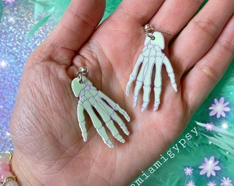 May include: A pair of silver skeleton hand earrings with a translucent white and purple iridescent finish. The earrings are held in a hand against a green feathery background with white flowers.