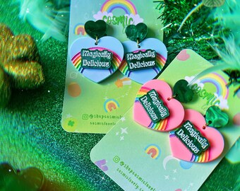 May include: A pair of heart-shaped earrings with a rainbow and the text "Magically Delicious" on each earring. The earrings are on a light blue and green background with a rainbow and stars.