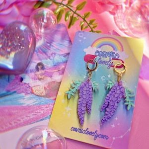 May include: A pair of purple wisteria earrings with gold hoops and green leaves. The earrings are displayed on a card with the text "cosmic lovely" and the website address "cosmiclovely.com".