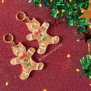 May include: Two gold gingerbread man-shaped keychains with white icing and red and green accents. The keychains are on a red glitter background with green tinsel and lights.