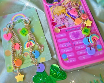 May include: A pink and clear phone case with a cartoon image of two girls. The case has a charm attached with a rainbow, hearts, stars, and a clover. The phone case has a keypad with numbers and letters. The text on the phone case reads "Beautiful girl".