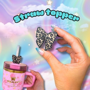 May include: A bow-shaped straw topper featuring a black and tan leopard print pattern. The text "Straw topper" is displayed above the item. The topper is shown both held in a hand and attached to the straw of a pink Stanley LoveShackFancy tumbler.