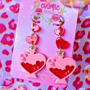 May include: Pink heart-shaped earrings with a red heart charm and a pink heart-shaped bottle filled with red glitter. The earrings are on a pink card with the text "cosmic lovely" and "cosmiclovely.com".