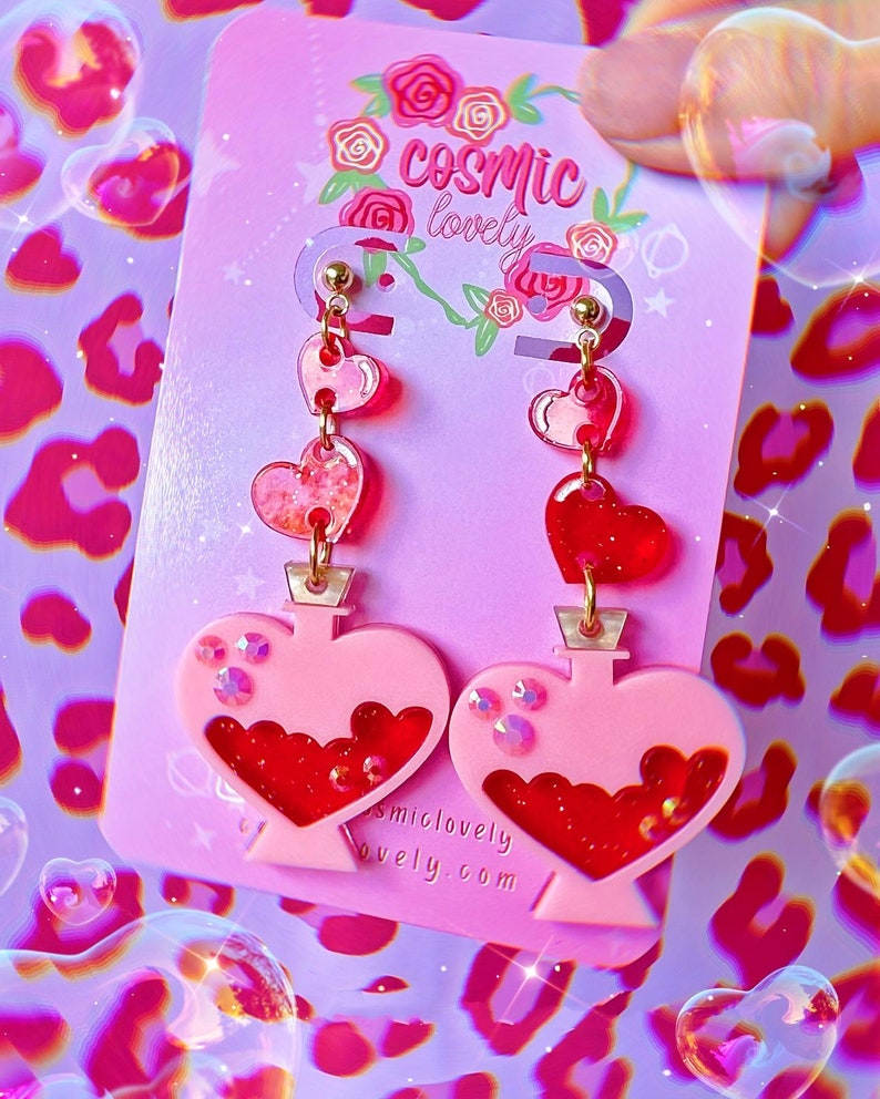 May include: Pink heart-shaped earrings with a red heart charm and a pink heart-shaped bottle filled with red glitter. The earrings are on a pink card with the text "cosmic lovely" and "cosmiclovely.com".