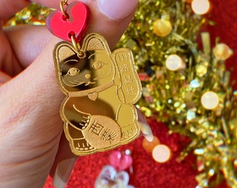 Peut inclure: Boucles d'oreilles en forme de Maneki-Neko, ou chat porte-bonheur, dorées avec un charme en forme de cœur rouge. Le chat tient une pièce d'or avec les caractères japonais "招福" (shohuku) signifiant "inviter la fortune".