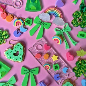 May include: Assortment of green, pink, and rainbow-colored resin earrings and charms. The earrings feature bows, hearts, rainbows, shamrocks, and a gumball machine. The charms are shaped like stars, hearts, and shamrocks.