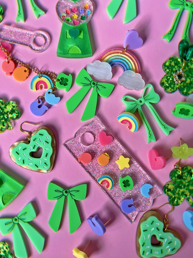 May include: Assortment of green, pink, and rainbow-colored resin earrings and charms. The earrings feature bows, hearts, rainbows, shamrocks, and a gumball machine. The charms are shaped like stars, hearts, and shamrocks.