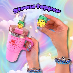 May include: A pink Stanley tumbler with a heart pattern and a yellow chick wearing a green frog hat straw topper. The text Straw topper is displayed at the top. A matching yellow chick straw topper is held separately in the frame.