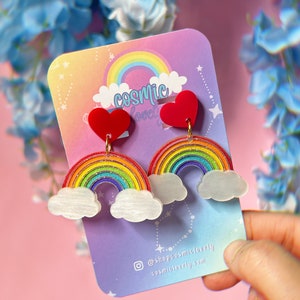 May include: Rainbow and heart shaped earrings with glitter accents. The earrings are on a white card with the text "cosmic lovely" and social media information.