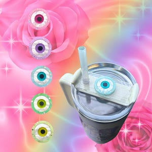 May include: A stainless steel tumbler with a white handle and a lid featuring a blue eye design. Several decorative eye-shaped accessories in pink, purple, blue, green, and yellow are also displayed. The background has a pink and rainbow gradient.
