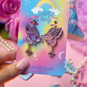 May include: A pair of purple iridescent bee earrings with gold accents and a purple gemstone dangle. The earrings are on a card with the text "cosmic lovely" and "cosmiclovely.com".