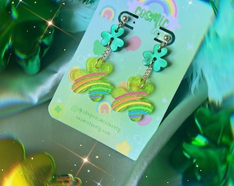 May include: Green shamrock earrings with a rainbow design and glitter. The earrings are on a white card with the text "Cosmic Lovely" and a rainbow.