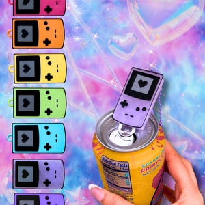 May include: A collection of seven handheld game console-shaped soda can tabs in pink, orange, yellow, green, blue, purple, and lavender. Each tab features a black screen with a heart icon, a directional pad, and buttons, attached to a yellow soda can.