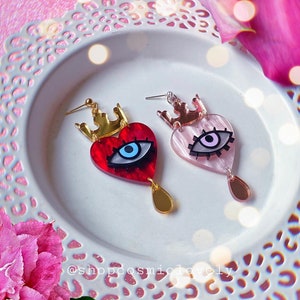 Puede incluir: Un par de pendientes con forma de coraz&oacute;n rojo y rosa con un dise&ntilde;o de ojo malvado. Cada coraz&oacute;n tiene una corona dorada en la parte superior y una l&aacute;grima dorada que cuelga debajo. Los pendientes est&aacute;n sobre un plato blanco con un fondo floral rosa.