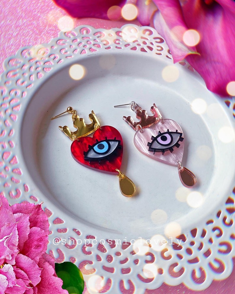 Puede incluir: Un par de pendientes con forma de coraz&oacute;n rojo y rosa con un dise&ntilde;o de ojo malvado. Cada coraz&oacute;n tiene una corona dorada en la parte superior y una l&aacute;grima dorada que cuelga debajo. Los pendientes est&aacute;n sobre un plato blanco con un fondo floral rosa.