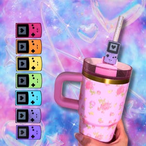 May include: A pink floral tumbler with a handle and a lid featuring a purple handheld gaming console straw topper. To the left, a vertical column displays six handheld gaming console icons in pink, orange, yellow, green, blue, and purple.