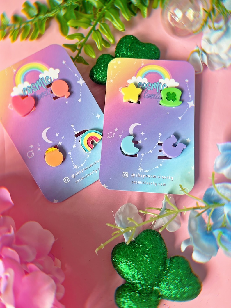 May include: Two sets of colorful, whimsical earrings on a pink background. The first set features a rainbow, a heart, a star, and a balloon. The second set features a shamrock, a star, a crescent moon, and a cloud. The cards are labeled "Cosmic Lovely" and include the social media handle @shopcosmiclovely and the website cosmiclovely.com.