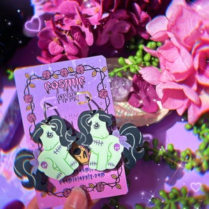 May include: Green and black zombie pony earrings with a pink bow and skull detail. The earrings are on a pink card with the text "cosmic lovely" and a website address.