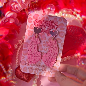 May include: Pink leopard print earrings with a pink heart charm. The earrings are on a pink and white card with the text "cosmic lovely" and the social media handle "@shopcosmiclovely" and website "cosmiclovely.com".