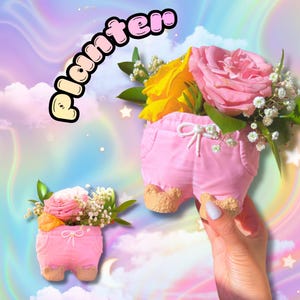May include: Pink planter shaped like shorts, filled with yellow and pink roses, white baby's breath, and green leaves. The word "Planter" is written in a bubble font. The planter has tan-colored feet.