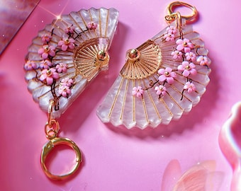 May include: A pair of white and gold fan-shaped keychains with pink cherry blossom accents. The keychains are on a pink background.