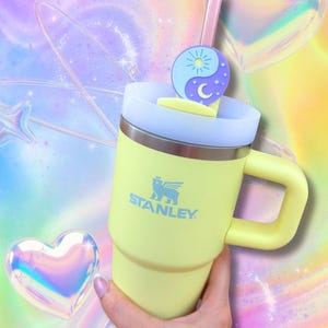 May include: A pale yellow Stanley tumbler with a handle, a light blue lid, and a pink straw. A circular yin-yang charm featuring a sun and moon design is attached to the straw. The Stanley logo is printed in light blue on the front of the cup.