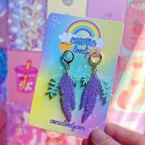 May include: Purple wisteria flower earrings with gold hoops and a green leaf design. The earrings are on a card with the text "cosmic lovely" and "cosmiclovely.com".
