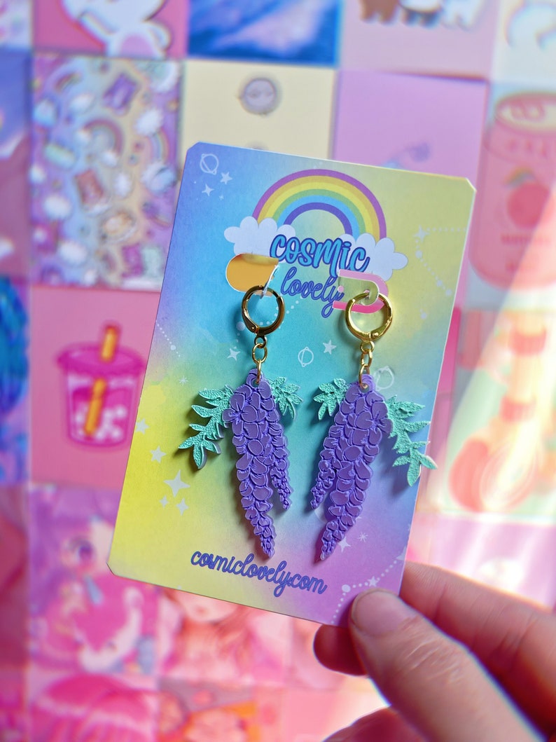 May include: Purple wisteria flower earrings with gold hoops and a green leaf design. The earrings are on a card with the text "cosmic lovely" and "cosmiclovely.com".