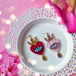 Puede incluir: Un par de pendientes en forma de coraz&oacute;n con una corona y un dise&ntilde;o de ojo. Un pendiente es rojo con un ojo azul, y el otro es rosa con un ojo morado. Los pendientes est&aacute;n en un plato blanco con un patr&oacute;n de encaje.