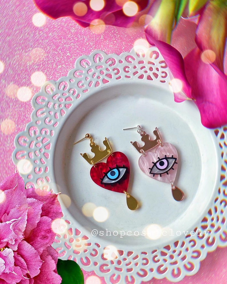 Puede incluir: Un par de pendientes en forma de coraz&oacute;n con una corona y un dise&ntilde;o de ojo. Un pendiente es rojo con un ojo azul, y el otro es rosa con un ojo morado. Los pendientes est&aacute;n en un plato blanco con un patr&oacute;n de encaje.