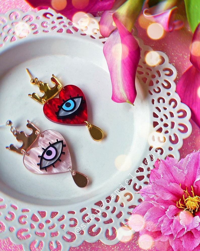 Puede incluir: Un par de pendientes en forma de coraz&oacute;n con una corona y un dise&ntilde;o de ojo. Los pendientes est&aacute;n hechos de acr&iacute;lico y tienen un acabado dorado. Un pendiente es rojo con un ojo azul, y el otro es rosa con un ojo morado.