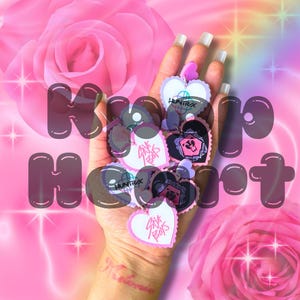 May include: A hand holding a collection of heart-shaped charms with various designs and text, including "PINK BOYS" and "HUNTRX". The background is a gradient of pink and purple with roses and the text "Momo Heart".