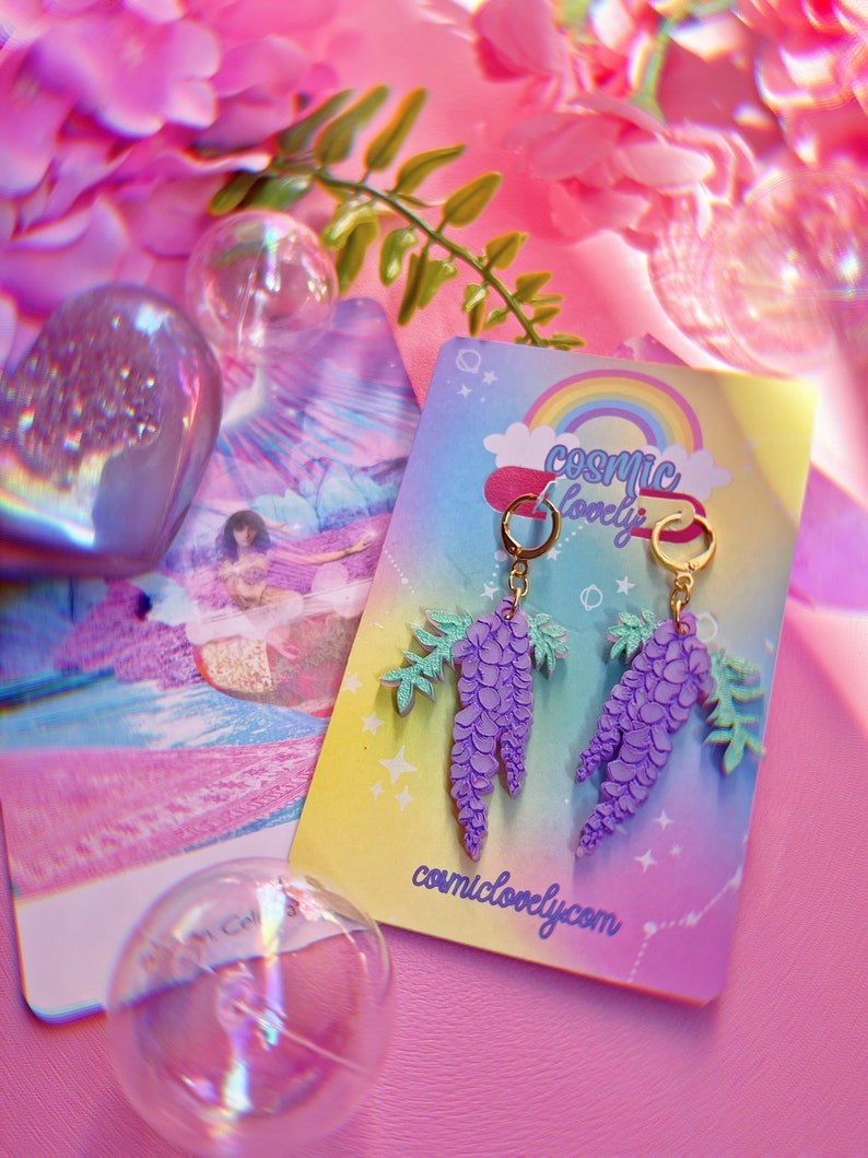 May include: A pair of purple wisteria earrings with gold hoops and green leaves. The earrings are displayed on a card with the text "cosmic lovely" and the website address "cosmiclovely.com".